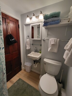 Un baño con inodoro, lavamanos y espejo. Hay toallas y un cubo de basura.