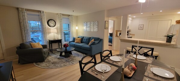Sala de estar con sofá azul, sillón oscuro, una mesa de centro y una zona de comedor para cuatro, con una araña.