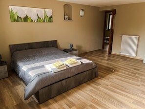 A bedroom with a large bed, wooden flooring, a nightstand, a lamp, a painting of flowers, a clock, and a radiator.
