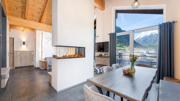 A modern kitchen with a large window, a dining area with a wooden table, and a fireplace. - 24 By Avenida Hotel and Residences Kaprun (Kaprun)