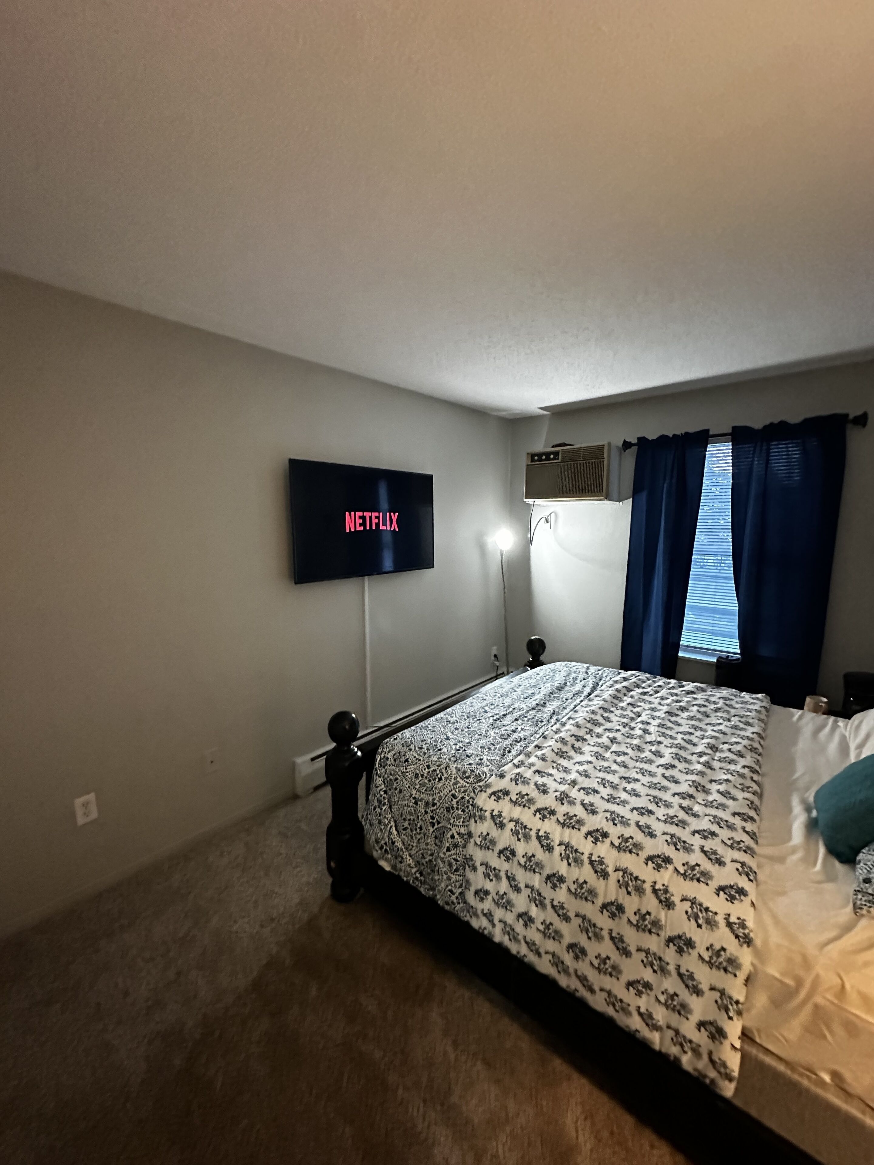 A bedroom with a bed, a wall-mounted TV displaying 'NETFLIX', a window with curtains, and an air conditioning unit.