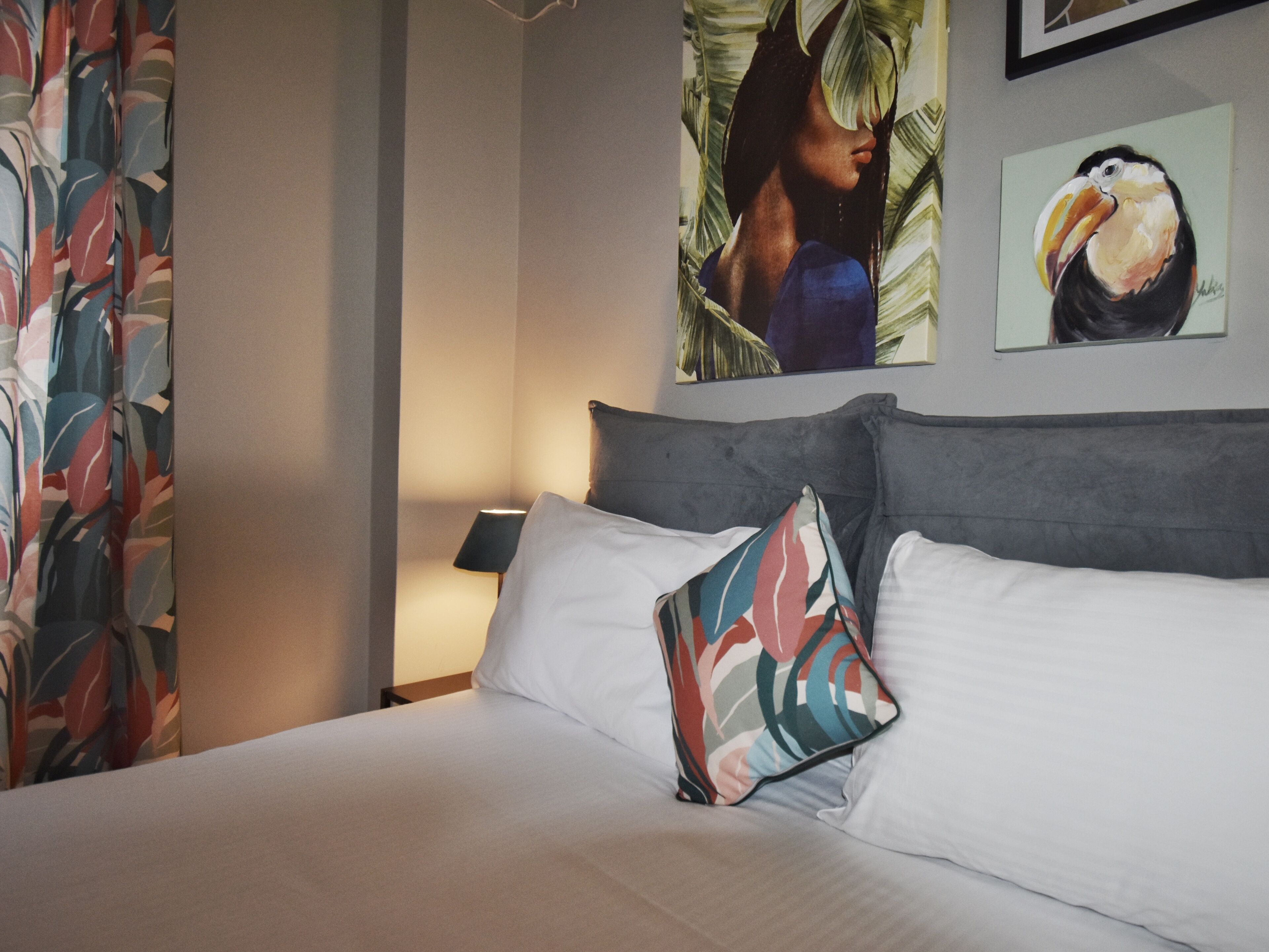 Una camera d'hotel con un letto, cuscini, un comodino, una lampada e due quadri appesi al muro.