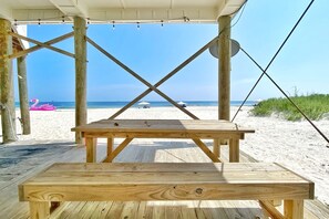 A wooden deck with a picnic table and a bench overlooking a beach with a pink flamingo float and umbrellas in the distance.