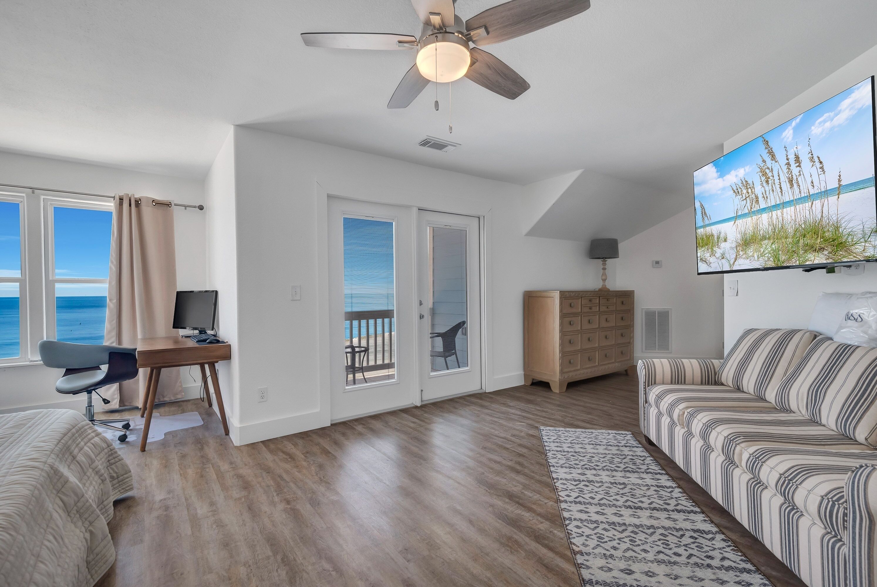 A spacious living room with a ceiling fan, a large TV, a sofa, a desk, and a view of the ocean through the windows.