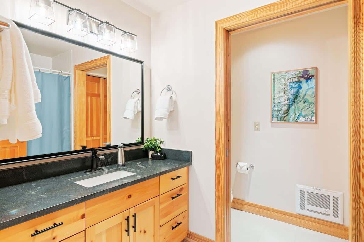 Une salle de bain avec un grand miroir, des meubles en bois, un plan de travail noir et une carte encadrée accrochée au mur.