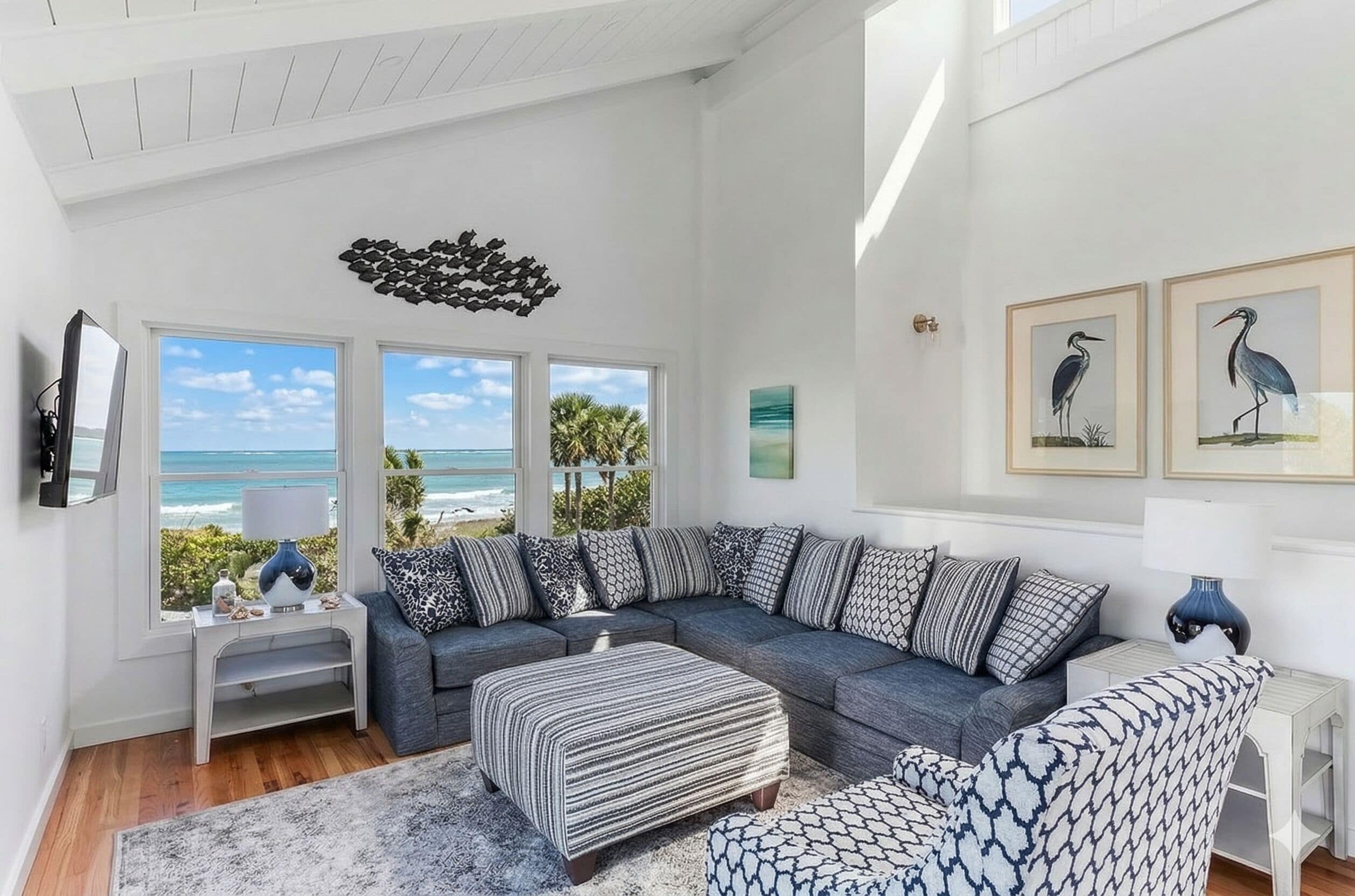 A modern living room with a grey sectional sofa, a patterned ottoman, a white coffee table, and a large window overlooking the ocean.