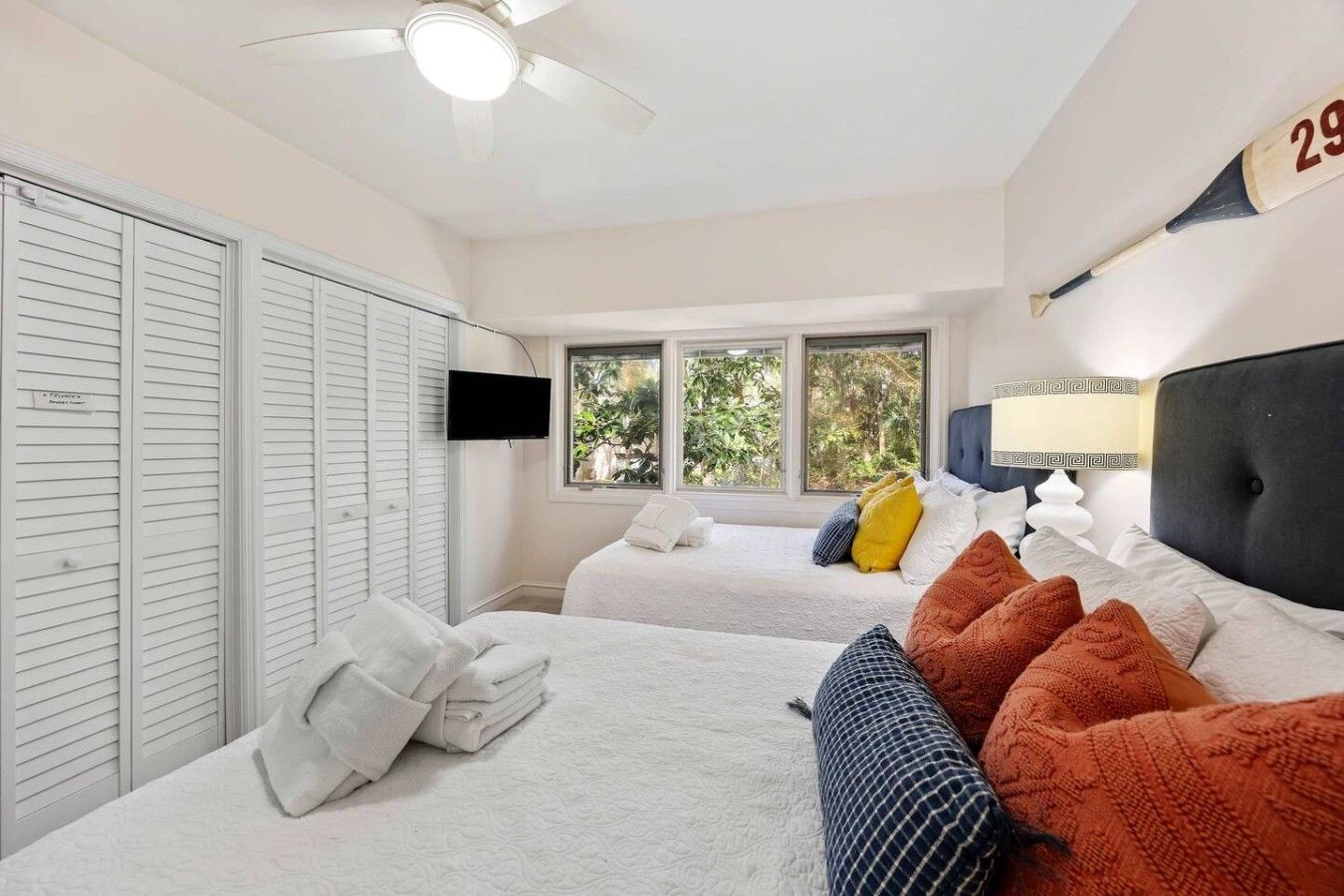 A bedroom with two beds, a ceiling fan, a TV, and a window with a view of greenery.