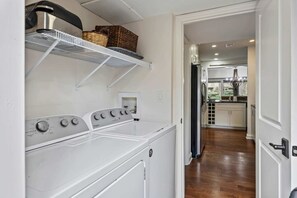 A laundry area with a front-loading washer and dryer, shelves above, and a view into a kitchen with wooden flooring.