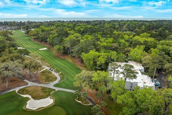 An aerial view of a golf course with a clubhouse, surrounded by dense trees and a clear sky.