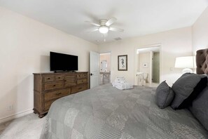 A bedroom with a bed, a dresser, a television, a ceiling fan, a bathroom visible through an open door, and a framed picture on the wall.