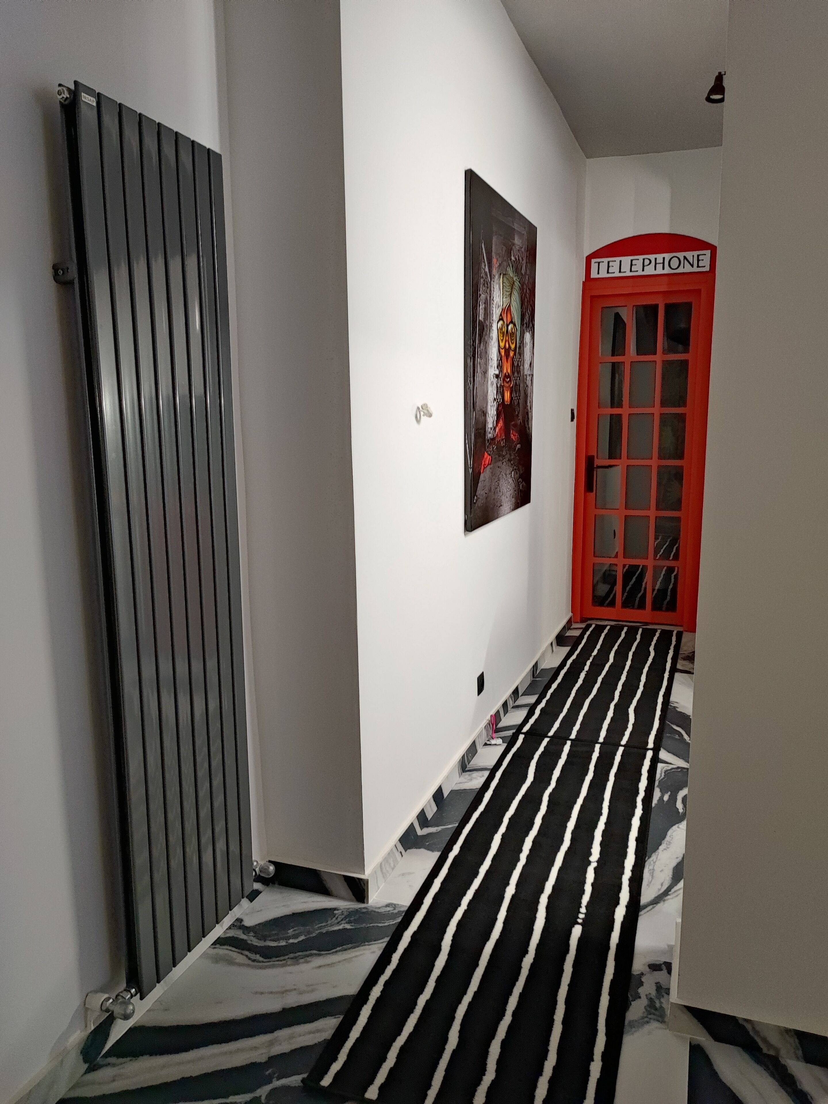 Un corridoio con tappeto a righe bianche e nere, un termosifone, un quadro e una porta rossa con la scritta 'TELEPHONE'.