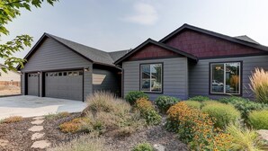Ein modernes, zweistöckiges Haus mit Garage, ein gepflegter Garten mit verschiedenen Pflanzen und Blumen, und ein klarer Himmel.