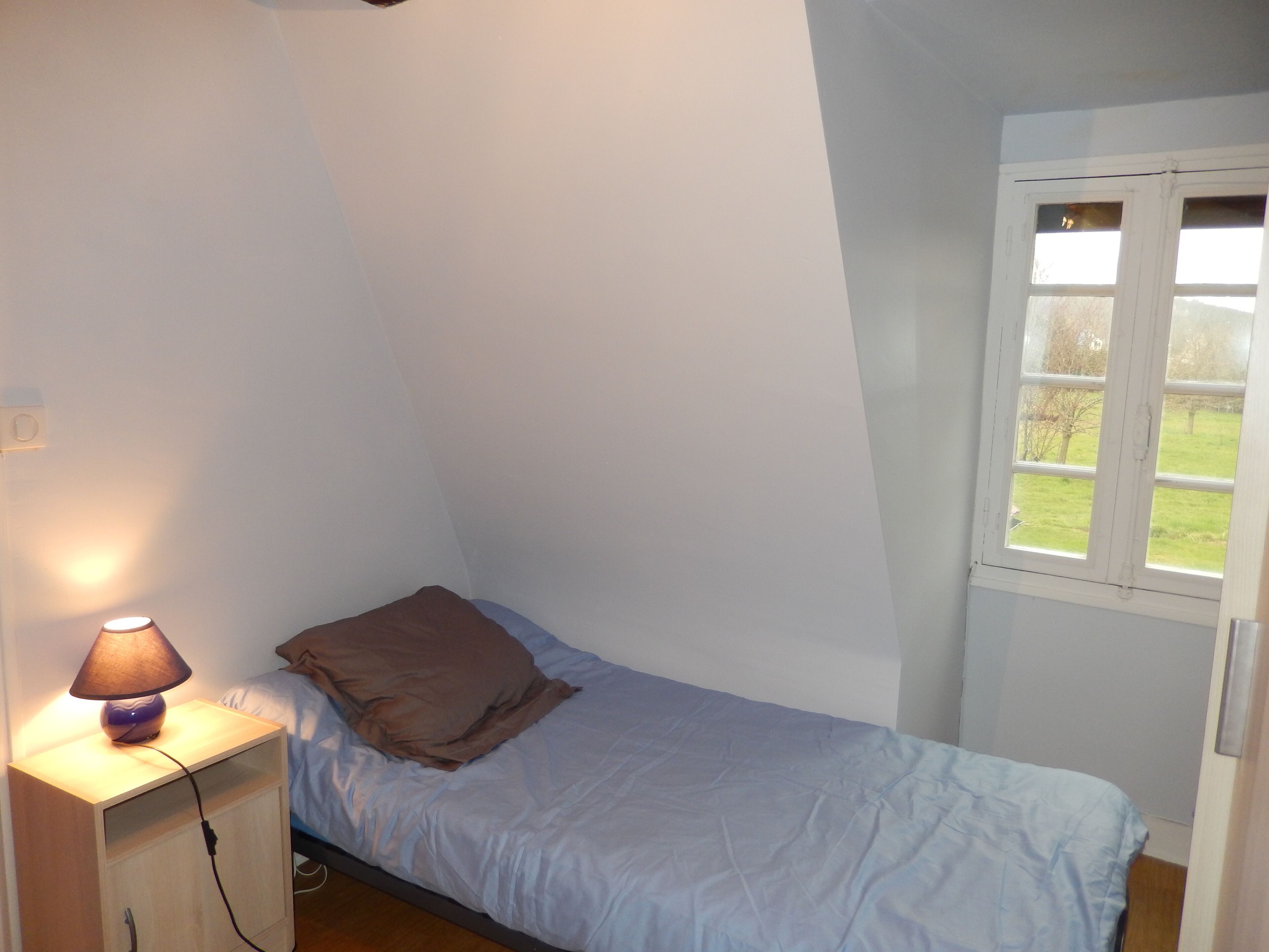 Ein kleines Zimmer mit einem Einzelbett, einem Nachttisch aus Holz, einer Lampe und einem Fenster mit Blick auf eine Rasenfläche.