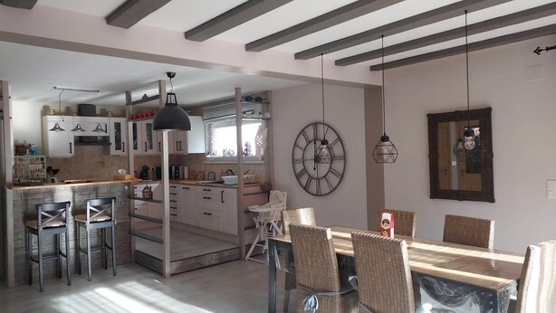 A modern kitchen with white cabinets, a dining area with wicker chairs, and a large wall clock. - Gîte Alsace Maison Avec Jardin Proche Strasbourg (Mutzig)