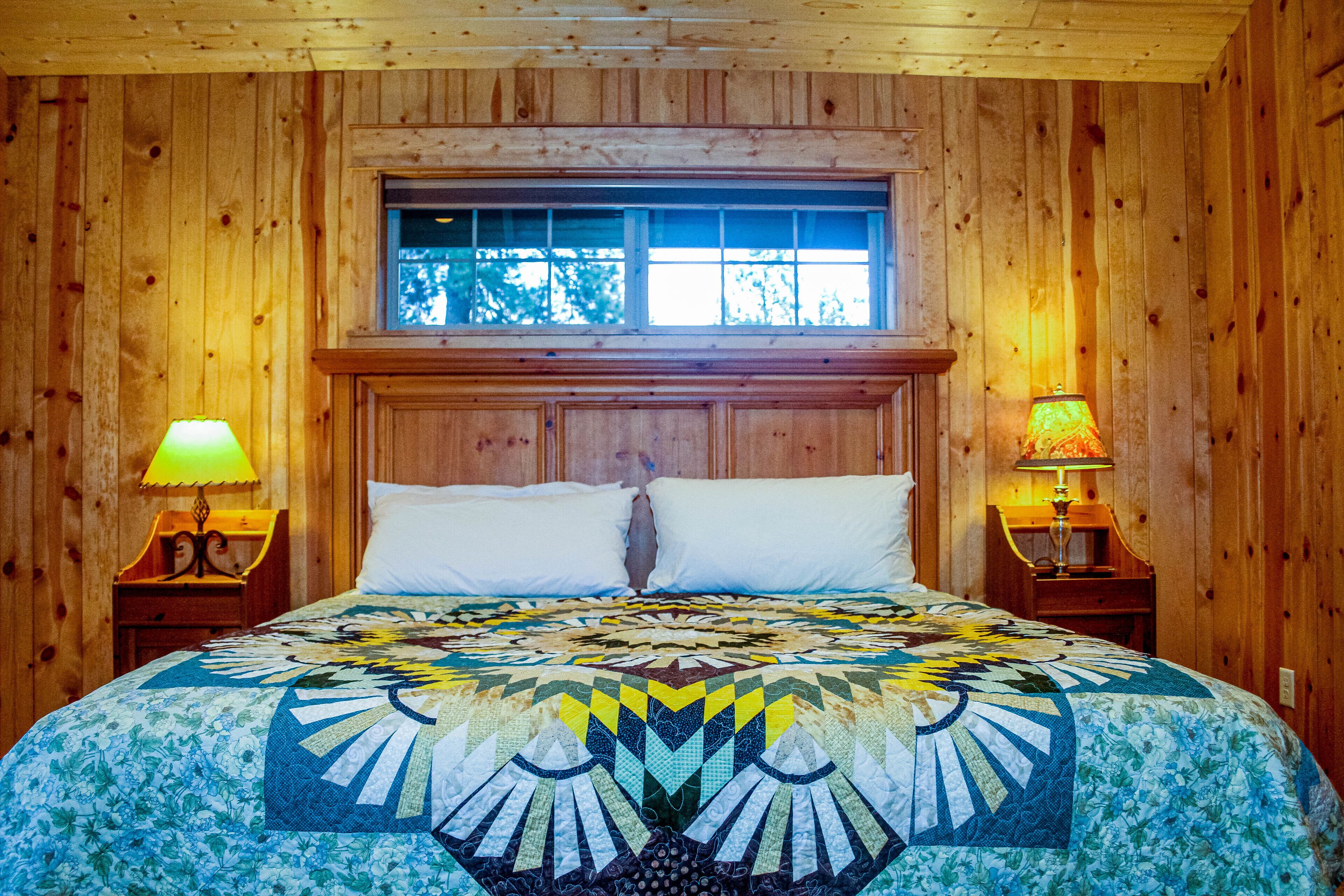 A wooden paneled room with a bed, two bedside lamps, and a window above the bed.