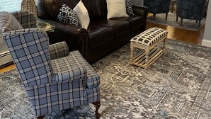 Una sala de estar con un sofá de cuero marrón oscuro, un sillón escocés y una alfombra estampada.