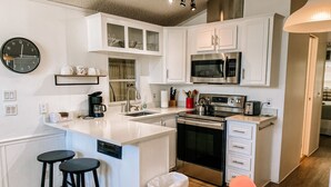 A modern kitchen with white cabinetry, a stainless steel stove, a microwave, and a sink. There are two black stools and a trash can. - Albey at the Beach (Sarasota)