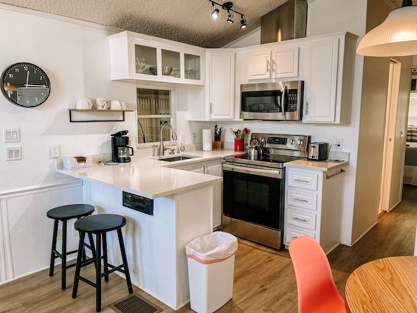 A modern kitchen with white cabinetry, a stainless steel stove, a microwave, and a sink. There are two black stools and a trash can.