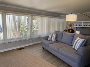 A living room with a grey sofa, striped pillows, a lamp, and a view through large windows with blinds.