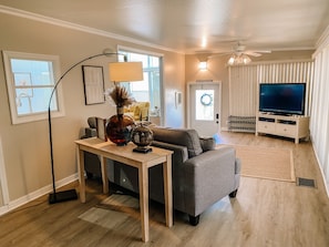 A living room with a grey sofa, a coffee table, a TV stand with a flat-screen TV, a floor lamp, and a door with a wreath.
