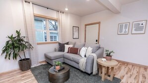 Un saló modern amb un sofà modular gris, una taula de centre rodona i una petita taula auxiliar de fusta. Hi ha una finestra gran amb vista a arbres, una planta en test i un quadre al mur.