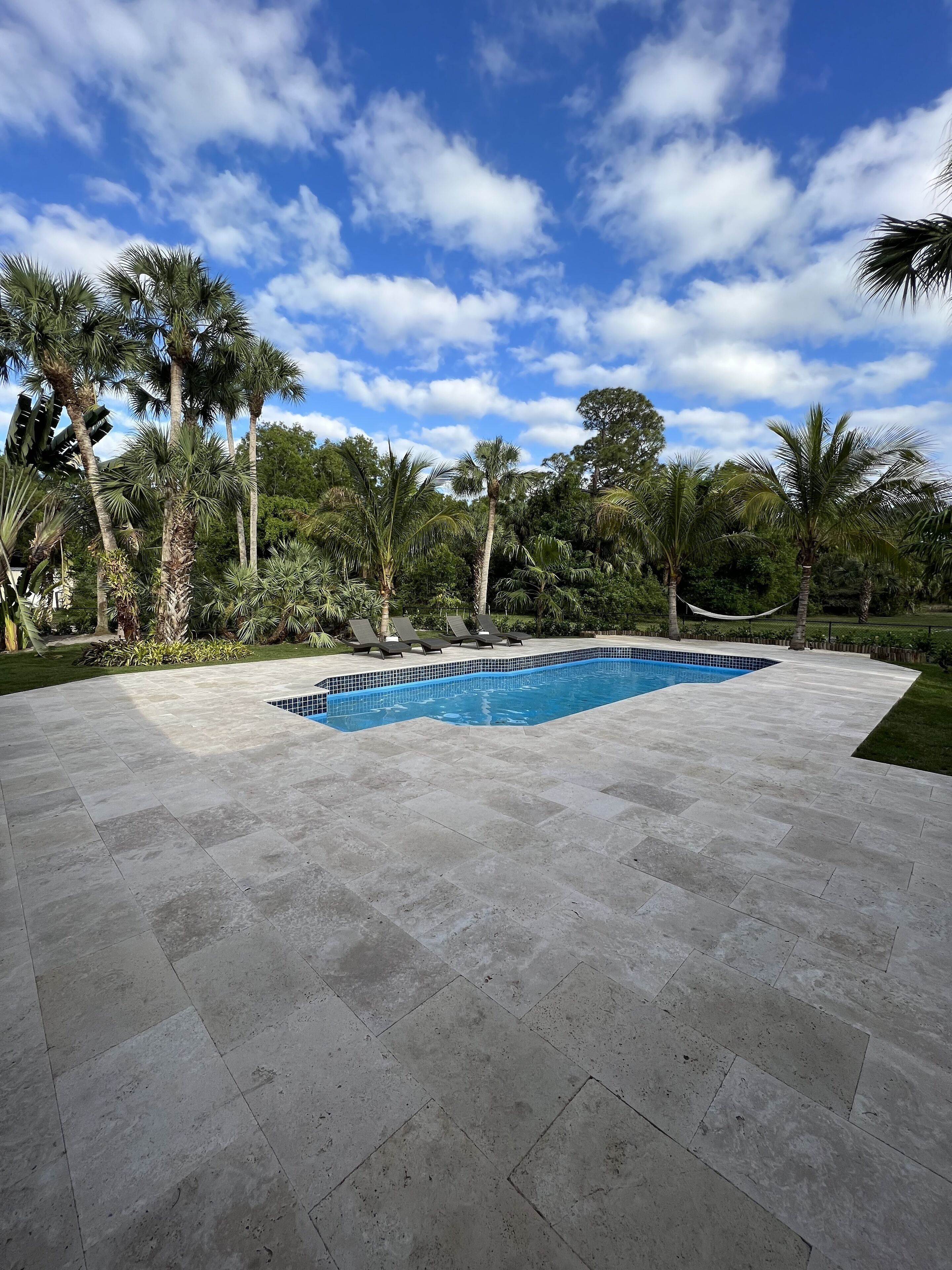A rectangular swimming pool with a tiled deck, surrounded by palm trees and a clear blue sky.
