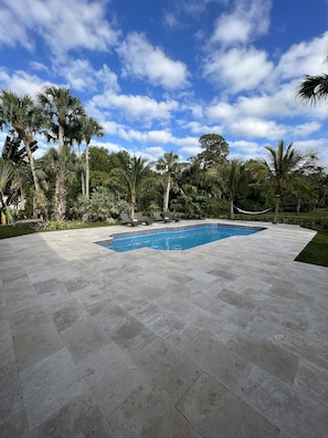 A rectangular swimming pool with a tiled deck, surrounded by palm trees and a clear blue sky.