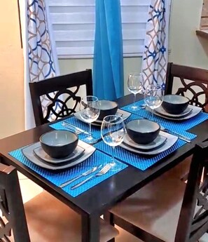 Een eettafel gedekt met zwarte kommen, borden en wijnglazen, omringd door stoelen met een blauwe tafelkleed.
