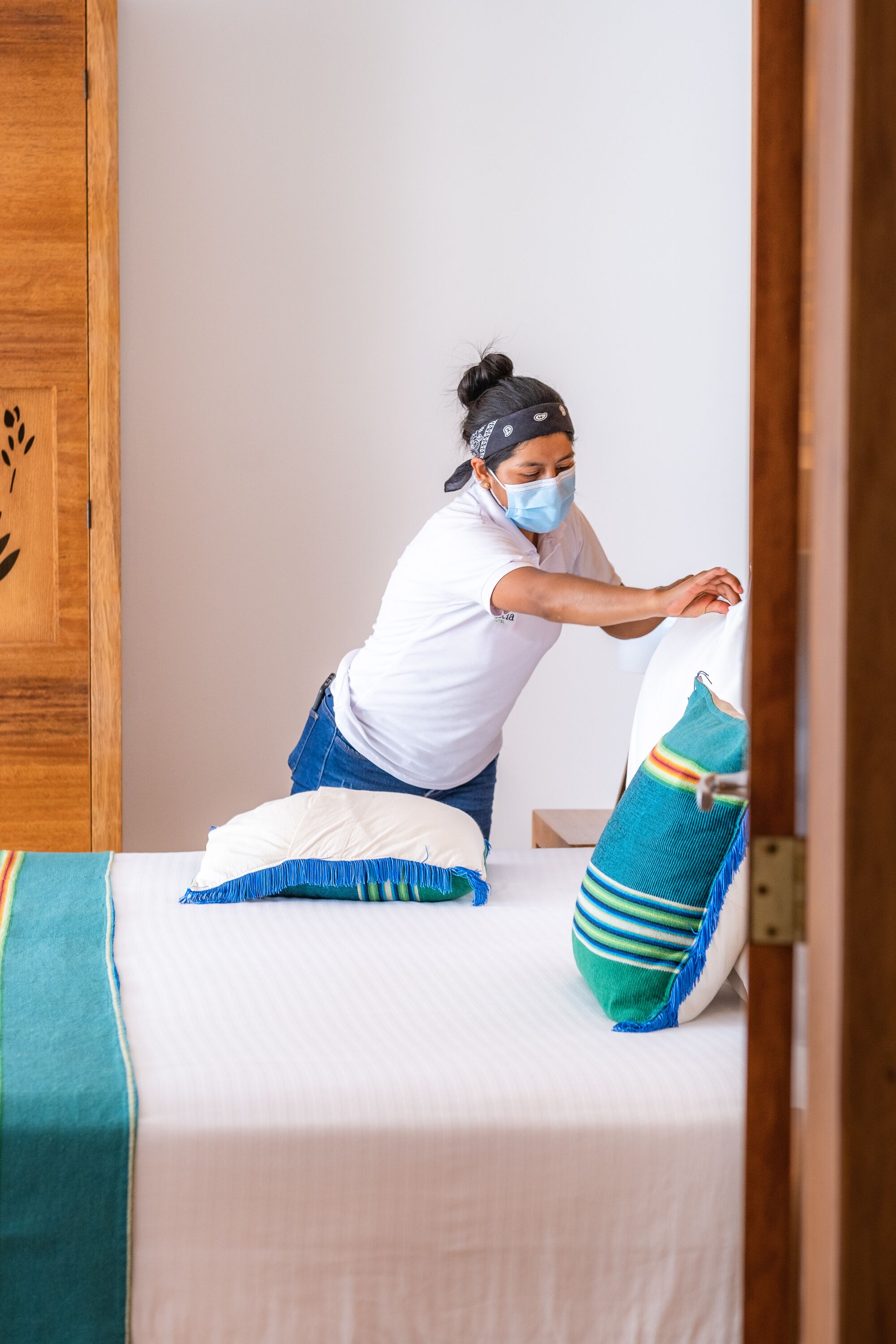 Une personne est en train de disposer un lit recouvert d’une couverture blanche et turquoise, avec des oreillers, et on aperçoit en arrière-plan une armoire en bois.