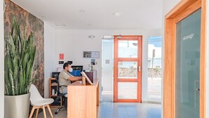 A reception desk with a person seated behind it, a potted plant, a white chair, and a wooden door leading outside. - Acacia Galapagos Boutique Hotel (Puerto Ayora)