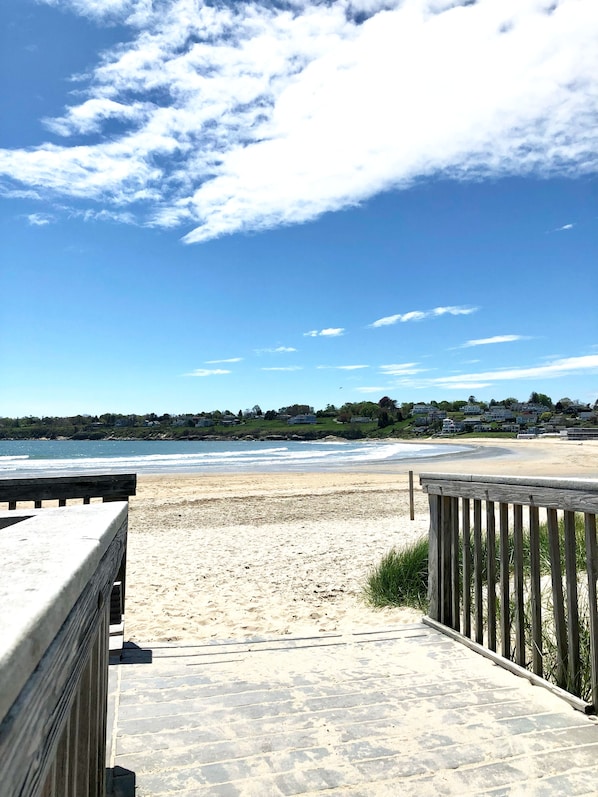 A beach with a wooden walkway leading to the shore, a clear sky, and houses in the background.