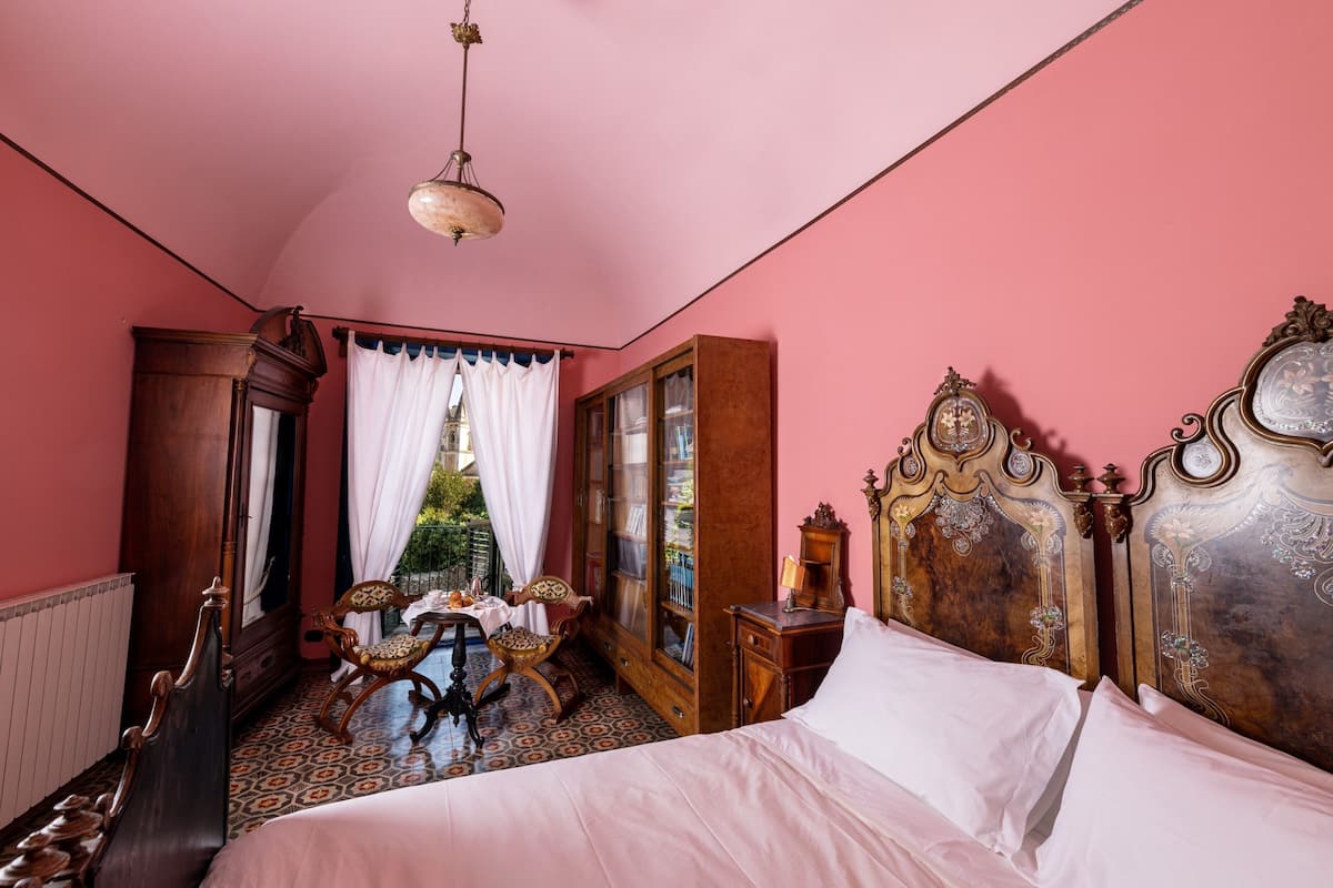 A room with a large bed, a wooden wardrobe, a small table with a chair, and a window with curtains.