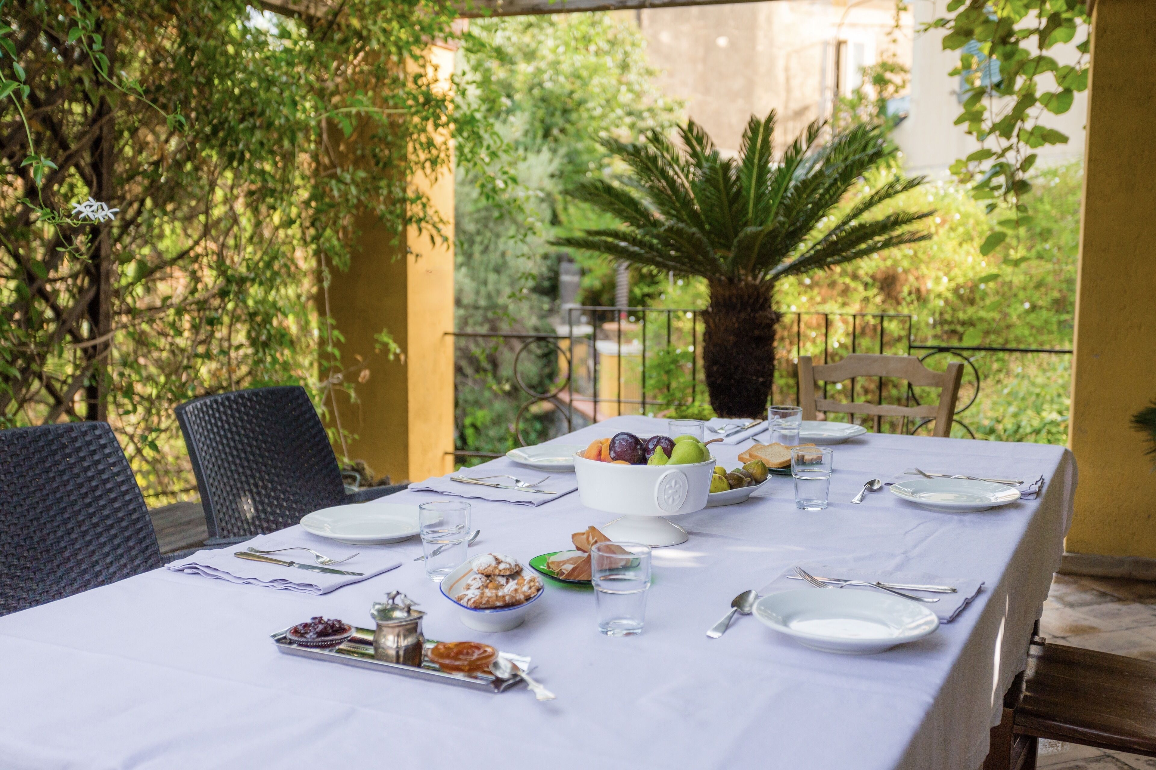 An outdoor dining table set with plates, glasses, and a bowl of fruit, surrounded by chairs and greenery.