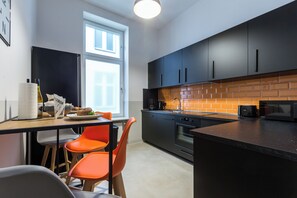 Een moderne keuken met zwarte kastjes, een achterwand van baksteen en een zwart aanrecht. Er staan twee oranje stoelen bij een witte tafel met een bord eten en een fles wijn.