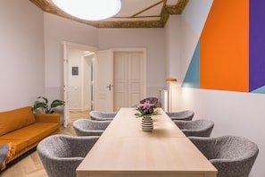 Een moderne kamer met een houten eettafel, grijs gestoffeerde stoelen, een gestreept vaasje met bloemen, een witte bank en een groot raam met uitzicht op de stad.