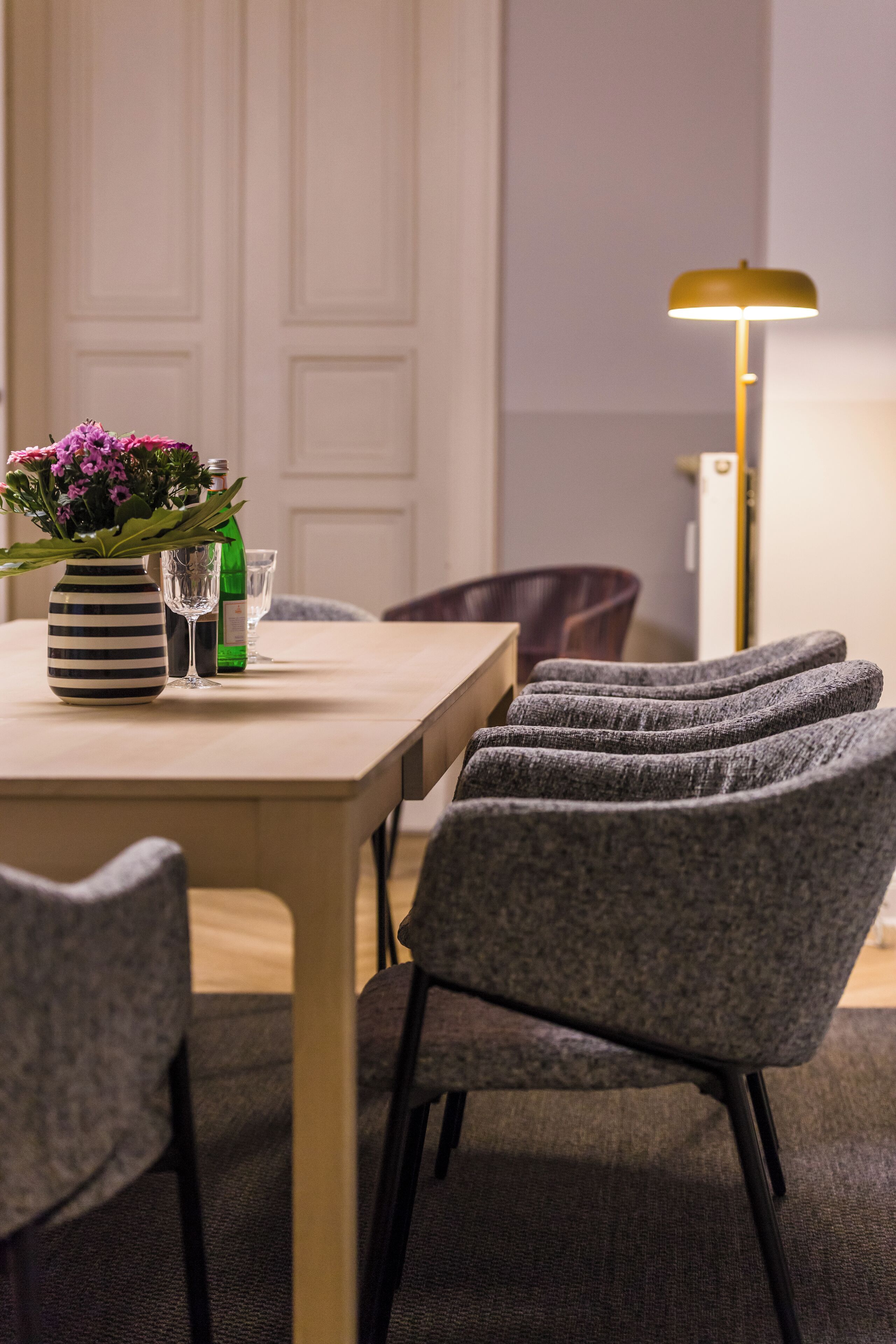 Un coin repas avec une table en bois, des chaises à revêtement gris, un vase rayé avec des fleurs, une bouteille verte et un grand lampadaire.