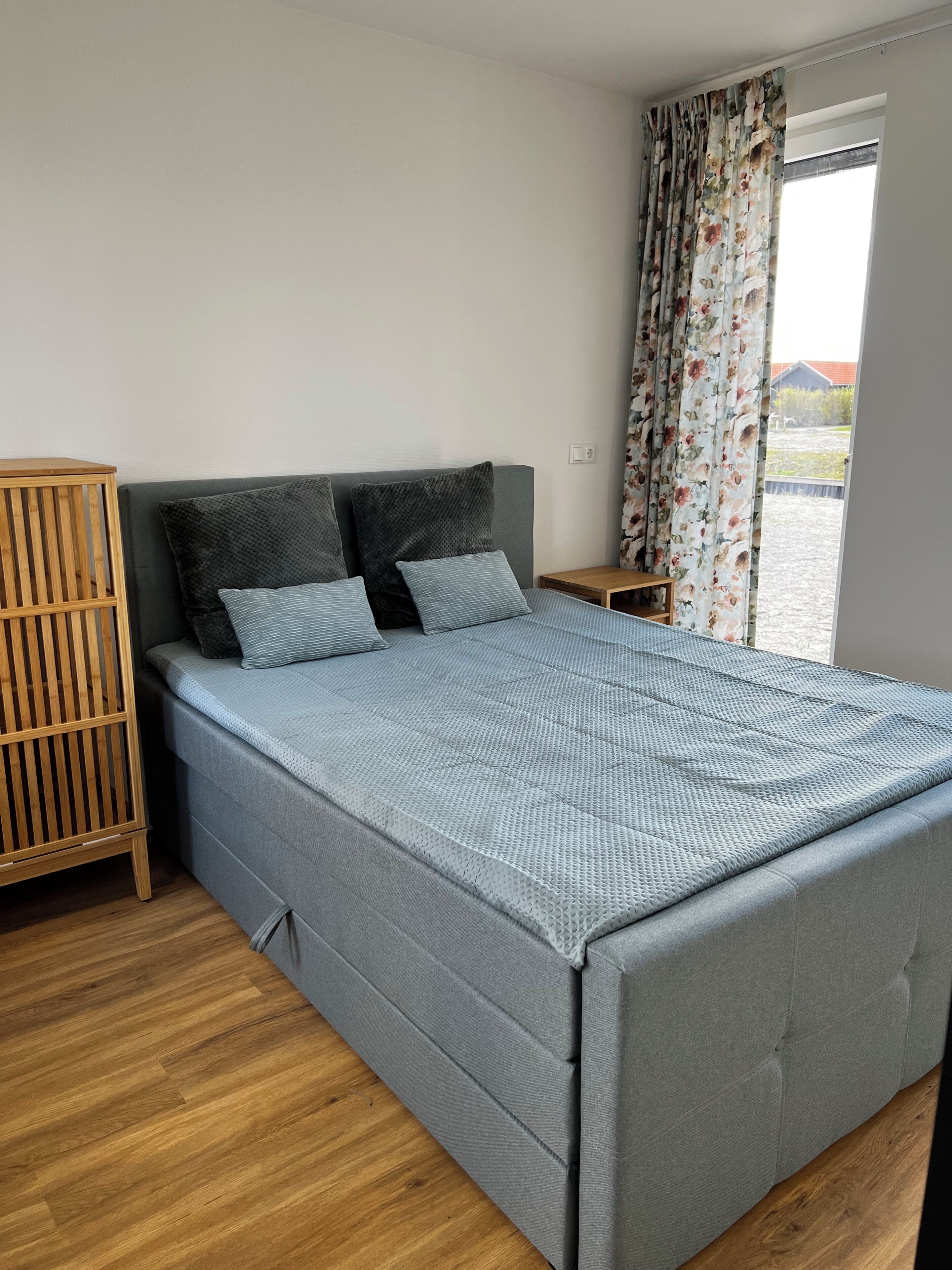 A bed with a grey upholstered headboard, a wooden nightstand, and a floral curtain.