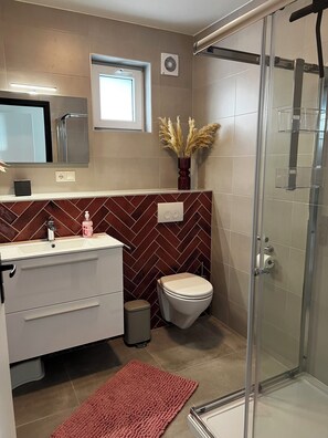 Un baño moderno con pared de diseño en patrón de espiga, cabina de ducha de vidrio y tocador con espejo y un florero de plumas de pájaro.