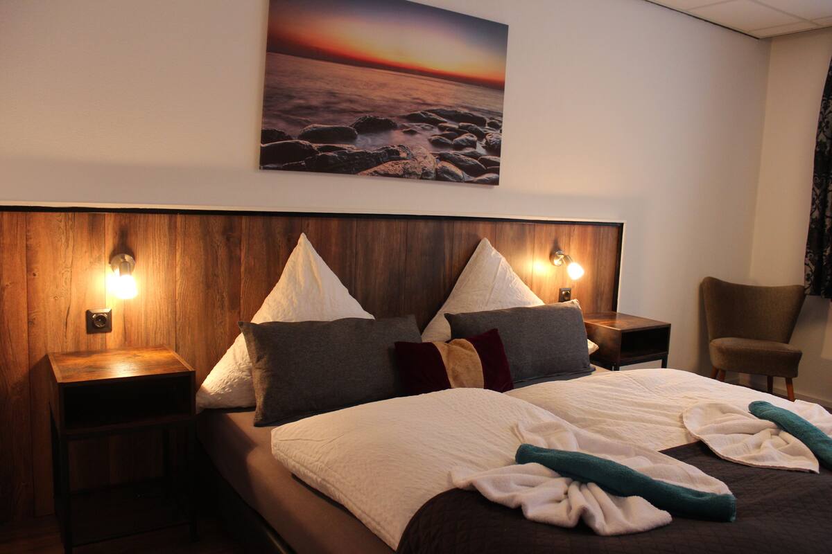A hotel room with a wooden headboard, a large bed with multiple pillows, a nightstand, a chair, and a painting of a sunset over the ocean.