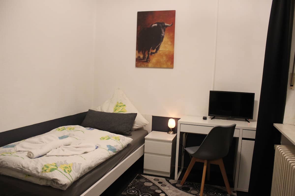 A bedroom with a bed, a desk, a chair, a TV, and a painting on the wall.