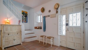 Una habitación con una escalera, un armario, dos taburetes, una puerta con detalles de vidrio y una pared con un cuadro y sombreros.