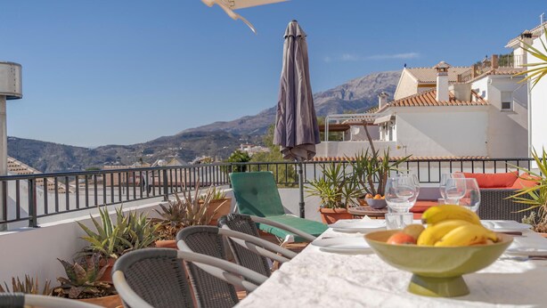 Una terraza con una mesa dispuesta para comer, plantas en macetas y vistas a las montañas.