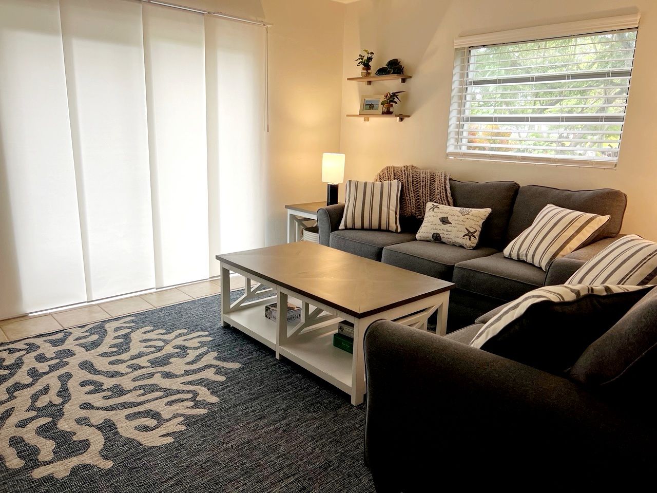 A living room with a sofa, a coffee table, a lamp, and a window with blinds.