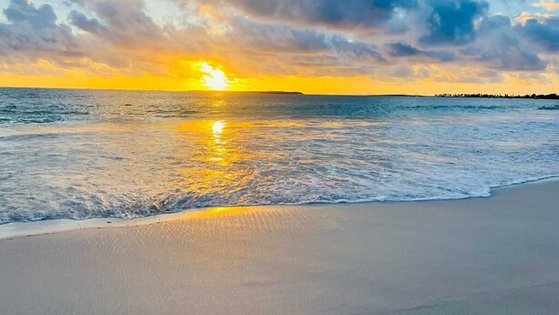 Una playa con un atardecer sobre el océano, olas tranquilas y un cielo despejado.
