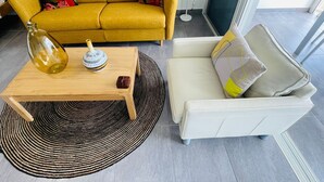 Una sala de estar con un sofá amarillo mostaza, un sillón blanco, una mesa de centro de madera con objetos decorativos y un tapete tejido en blanco y negro.