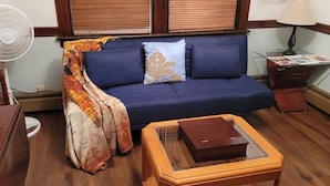 Une pièce avec un canapé bleu, une table d’appoint en bois, une petite table en bois sur laquelle se trouve une boîte, un ventilateur, une lampe et une fenêtre avec des stores.