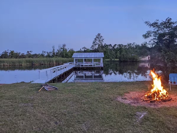 A dock extending into a calm body of water, a small wooden structure, and a campfire on the shore.