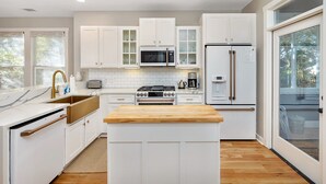 Uma cozinha moderna com armários brancos, uma ilha central, eletrodomésticos de aço inoxidável e uma grande janela com vista para o exterior.