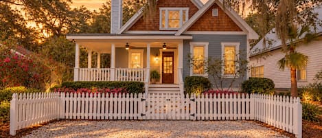 A two-story house with a porch, a white picket fence, and a gravel driveway.