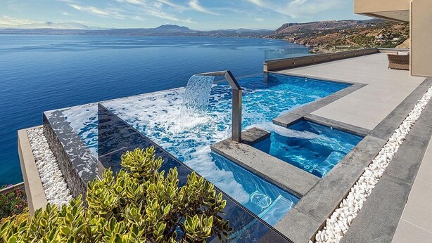 A modern infinity pool with a waterfall feature, surrounded by stone steps and plants, overlooking a scenic coastal view. - Sparkling ocean views with heated pool (Rodhia)